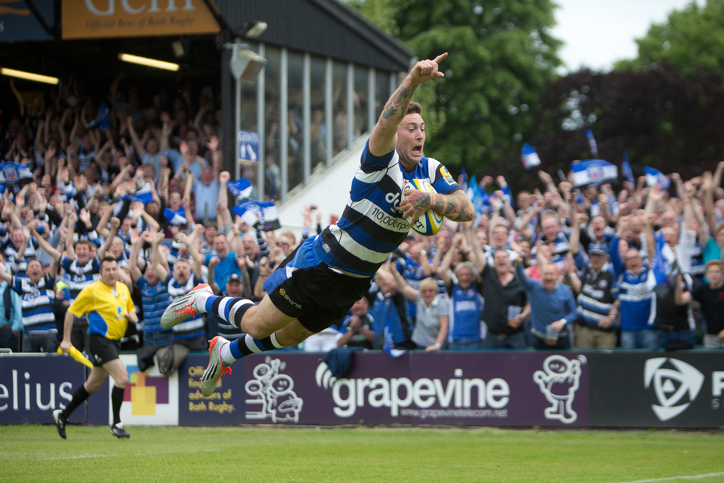 A Bath Rugby player photographed mid action with the Grapevine pitch side banner visible in the background.